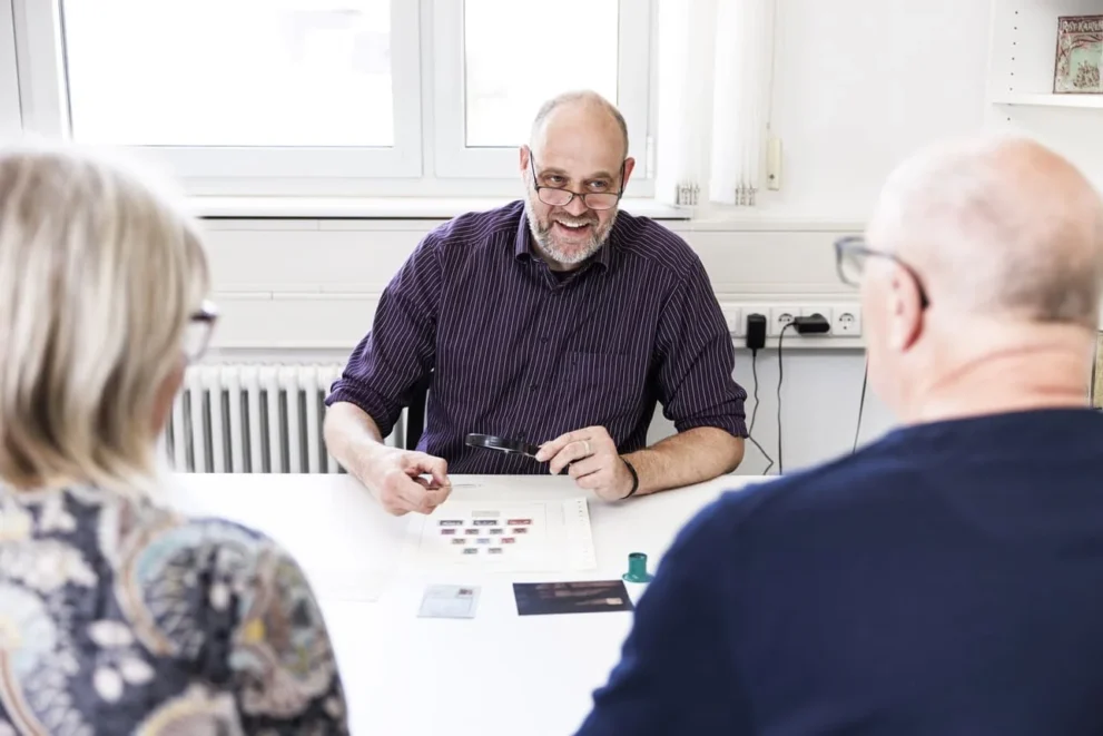 Mann mit Brille hält eine Briefmarke und eine Lupe in der Hand. Auf dem Tisch vor ihm liegen weitere Briefmarken.